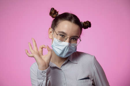 Modern young teen girl in protective medical facial mask showing okay gesture on pink studio background with copy spaceの写真素材