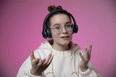 Bewildered young teen girl in headphones and glasses spread empty hands. Excited female in earphones choosing option on green studio background with copy space for ad content.の写真素材
