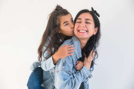 Little kid daughter kissing smiling mum in cheek, hugging behind. Mothers Day, happy motherhood conceptの写真素材