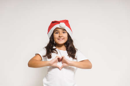 Smiling child girl in christmas santa hat showing heart gesture, congratulating with xmas holiday. Love, care and trustの写真素材