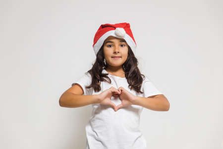 Smiling child girl in christmas santa hat showing heart gesture, congratulating with xmas holiday. Love, care and trustの写真素材