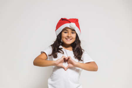 Smiling child girl in christmas santa hat showing heart gesture, congratulating with xmas holiday. Love, care and trustの写真素材