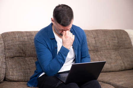 Overworked upset man massaging nose bridge, suffer headache, eyestrain after long working with laptop, sitting on couchの写真素材