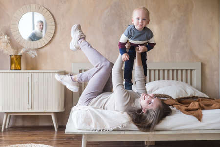 Joyful young mother hold son toddler imitate plane fly having fun play in funny game lying on bed in cozy homeの写真素材