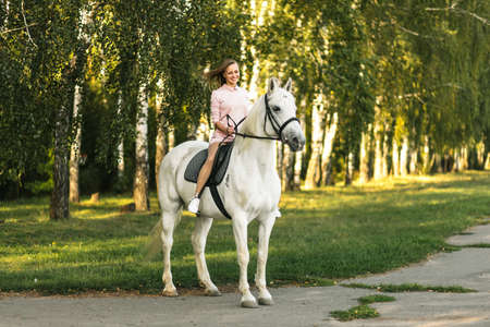 Happy young girl practicing horseback riding on thoroughbred white horse outdoors. Equestrianism school advertisingの写真素材