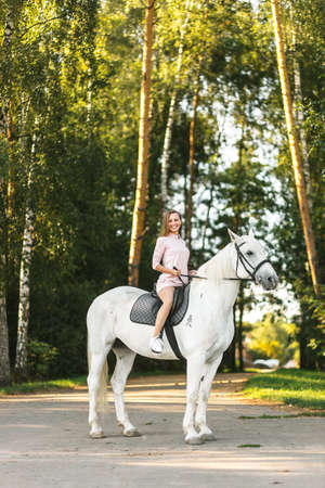 Happy young girl practicing horseback riding on thoroughbred white horse outdoors. Equestrianism school advertisingの写真素材