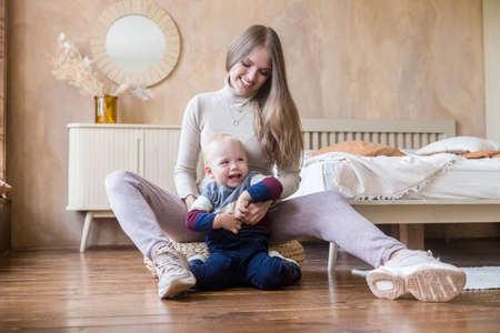 Happy young mother and little son having fun, tickling, sitting on wooden warm floor at home bedroom togetherの写真素材