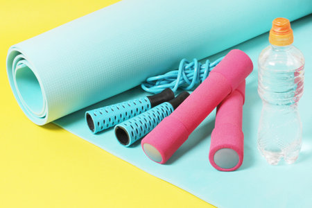 Fitness items jump rope, dumbbells, bottle of water and exercise mat on a yellow backgroundの写真素材