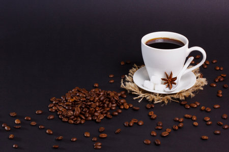 A white cup of black coffee sits on a saucer and jute mat with sugar cubes and star anise. Roasted coffee beans are scattered on a dark surface around it.の写真素材