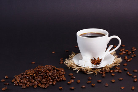 A white mug filled with black coffee sits on a saucer with sugar cubes and star anise. Coffee beans are scattered around the cup and a pile sits in the corner on a dark background.の写真素材