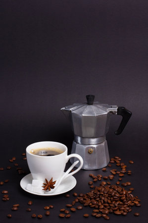 A cup of dark coffee sits next to a moka pot. Coffee beans are scattered around. Sugar cubes and star anise sit on the saucer. A white spoon rests on the saucer.の写真素材