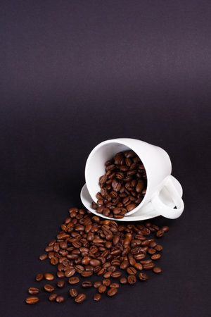 Roasted coffee beans overflow from a tipped over white cup with a saucer, spilling onto the dark, plain background. Focus on the textures and the rich color of the beans.の写真素材