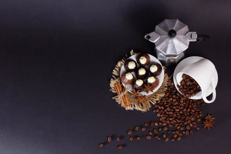 Coffee beans have spilled from a white cup onto a dark surface near a moka pot and a plate of chocolate candies. Cinnamon sticks and star anise add to the sweet treat arrangement.の写真素材