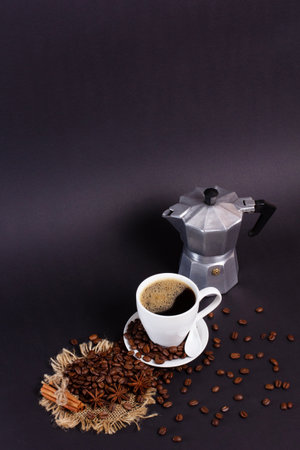 Still life features a cup of steaming coffee, an espresso maker, roasted coffee beans, star anise, and cinnamon sticks arranged on a dark surface.の写真素材