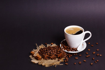 A white cup filled with black coffee sits on a saucer next to roasted beans, star anise, and cinnamon sticks. The ingredients are placed on a burlap surface against a dark background.の写真素材
