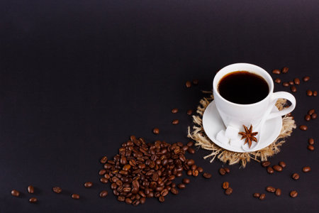 A full white cup of coffee sits on a saucer with sugar cubes and star anise. Coffee beans are scattered around the cup. The table is black.の写真素材