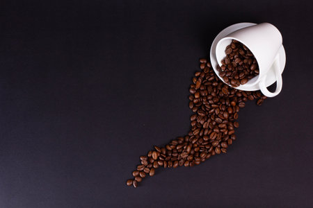 A white coffee cup rests on its side on a dark surface. Roasted coffee beans have spilled from the cup, creating a curved line or path across the dark background.の写真素材