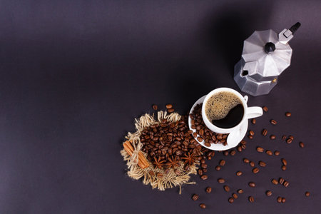 A cup of coffee sits next to an old moka pot, with surrounding coffee beans, cinnamon sticks, and star anise on a burlap base placed on a dark surface.の写真素材