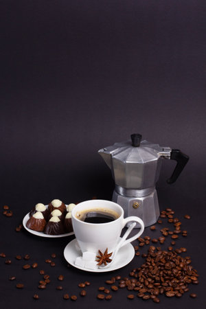 Dark coffee is served in a white cup with sugar cubes and star anise alongside chocolate candies on a white plate and a silver moka pot. Roasted coffee beans are scattered around.の写真素材