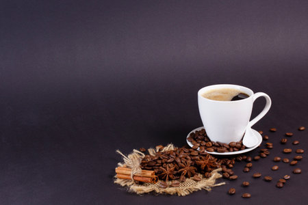 A cup of coffee sits on a saucer surrounded by coffee beans. Bundled cinnamon sticks and star anise rest on burlap, placed near the cup. The background is solid black.の写真素材