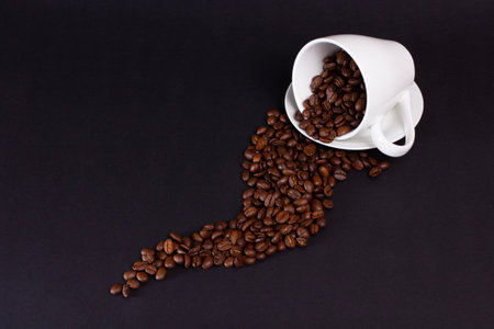 Coffee beans spilling from an overturned white coffee cup on a dark background. Beans are cascading and scattering, showing their roasted color and texture.の写真素材