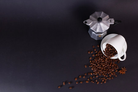 A white cup spills dark roasted coffee beans onto a black surface. A silver moka pot sits nearby, and an anise star is also present on the surface.の写真素材