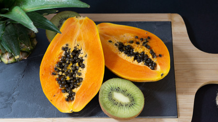 The papaya is cut into two halves, the stand is a stone board with a wooden frame, near the Papay there is a cut off top of the pineapple, the background is blackの写真素材