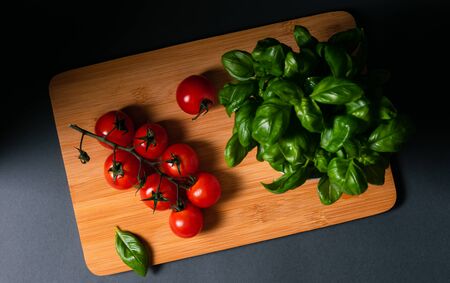 still life, a branch of cherry tomatoes with a bush of basil leaves on a wooden board on a black...
