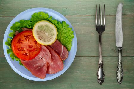 ham with salad, tomato and lemon on light blue plate. ham on green wooden background .ham wth fork and knife top viewの写真素材
