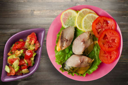 smoked mackerel on pink plate witn vegetable salad top view. mackerel with lemon and tomato on black wooden backgroundの写真素材