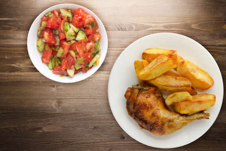 Chicken thigh with french fries on a brown wooden background. chicken thigh on a white plate with vegetable salad. rustic foodの写真素材