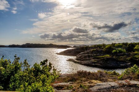 Evening sun over Flekkeroya. Seascape with rocky shores, cloudy skies and the bay, reflecting the...