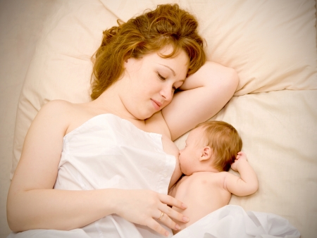 Young redhead mother is breast feeding a newborn baby and dreaming about the future  They are lying on the bed hidden by a sheet の写真素材