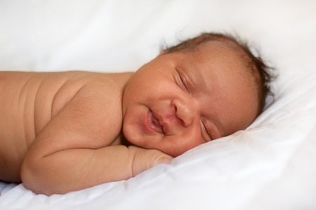 Mulatto newborn baby girl sleeping and smiling with his dreams. Black baby on white bed.の写真素材