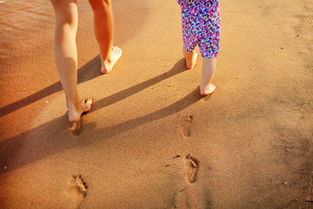 Mother and baby walking along the beach. Legs, feet and footprints on the sand. Summer vacation.の写真素材