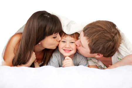 Happy parents kissing child in bed covered with a blanket. Good morning! Isolated on white background.の写真素材