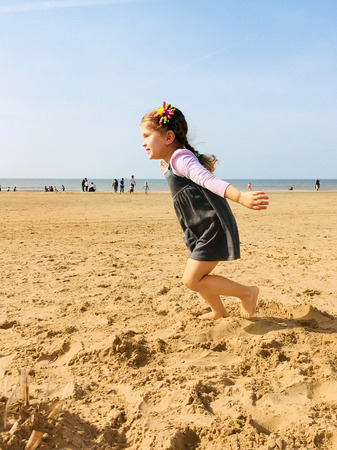 A small happy little girl runs barefoot along the beach towards the sun. A child runs, arms spread out like wings. Child happiness concept.の写真素材