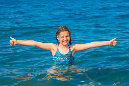 Cheerful girl swims in the sea. Child shows thumbs up sign. Smiling girl likes the sea and the beach. Happy summer holiday, summer vacation concept. Summer fun.の写真素材