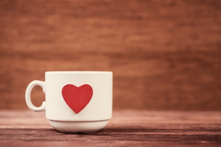 White cup with red heart shape over vintage defocused wooden background