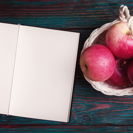 Open blank notebook and red apples in basket over rustic wooden background.の写真素材