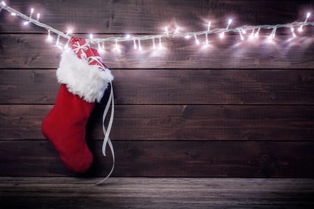 Red Santa stocking with gift boxes hanging on glowing garland over rustic wooden