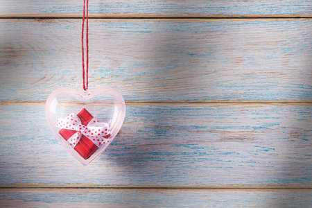Red gift box in transparent heart shaped container hanging over wooden background. Valentineâs day conceptual greetingの写真素材