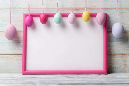 White blank card in pink frame and decorative Easter eggs over wooden background