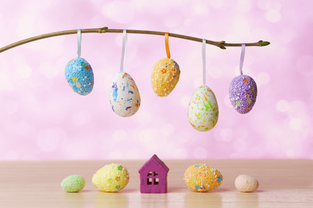 A festive Easter setting featuring colorful eggs hanging from a branch and miniature pastel houses set against a rustic blue background. Easter holiday conceptの写真素材