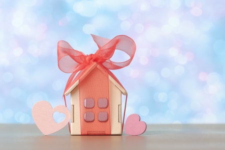 A miniature house wrapped in a red ribbon and bow, flanked by pink hearts, symbolizing love and celebration in property gifting or real estate investmentの写真素材