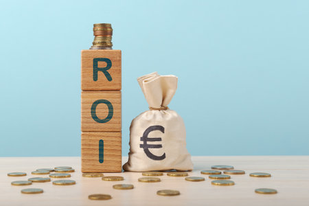 Wooden blocks spelling out ROI stacked beside a sack of Euros, symbolizing return on investment, on a table scattered with coins against a pale blue backgroundの写真素材