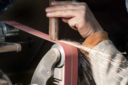 Blade sharpening creating on a belt grinder process. Knife making master hands close up.