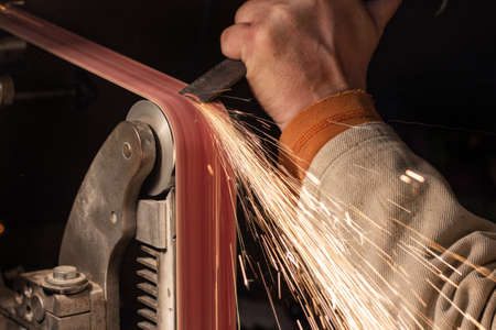 Blade sharpening creating on a belt grinder process. Knife making master hands close up.の写真素材