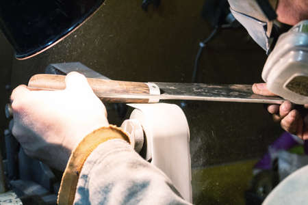 Blade sharpening creating on a belt grinder process. Knife making master hands close up.