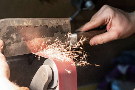 Blade sharpening creating on a belt grinder process. Knife making master hands close up.